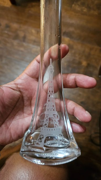 a vase that is eye shaped at the bottom and tapers. on one side is an image of the Eiffel Tower and the word bonjour. on the other side is t